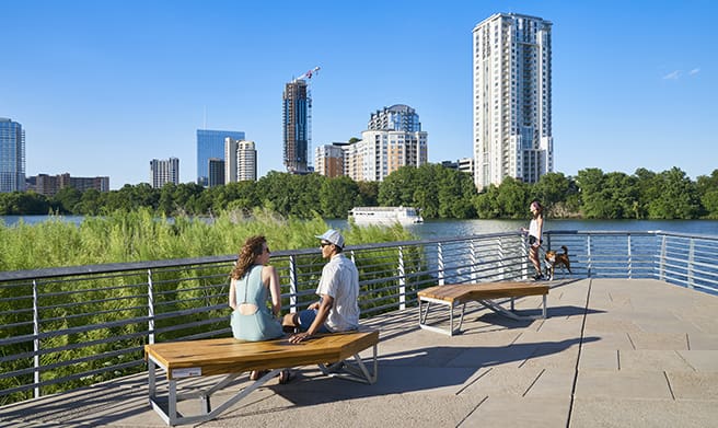 A woman and a man sit on a wooden bench on the Boardwalk with tall buildings and Lady Bird Lake in the background. 